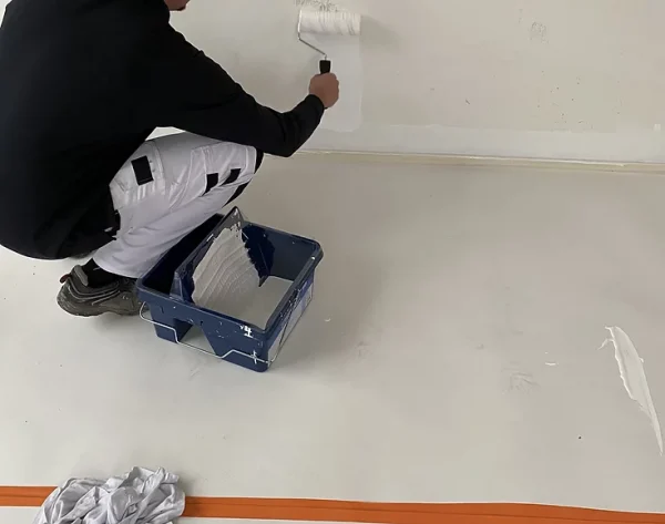 Mężczyzna maluje białą ścianę wałkiem stojąc na materiałach ochrony powierzchni