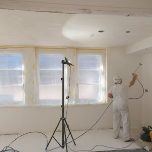 Mężczyzna maluje ścianę natryskowo w pokoju przed oknem zabezpieczonym folią
