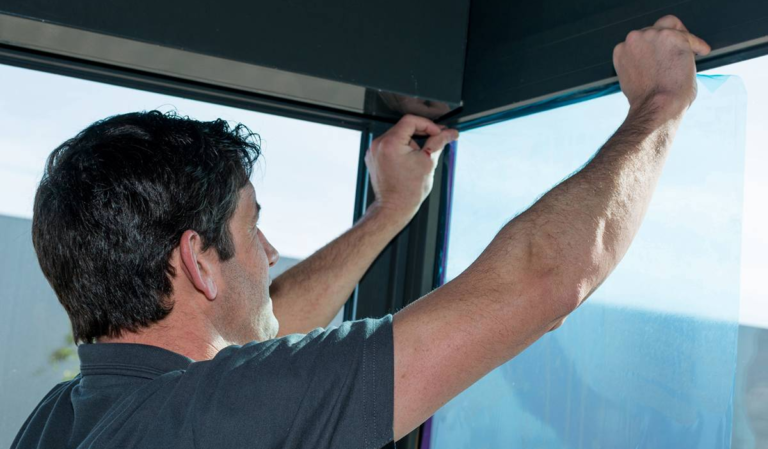 Mężczyzna przykleja niebieską folię ochronną na szybę okna.